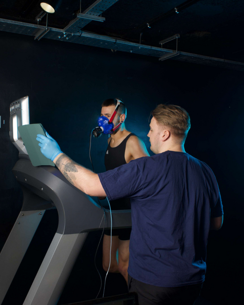 Person using a treadmill with another person wearing a mask and breathing apparatus, in a dark room.