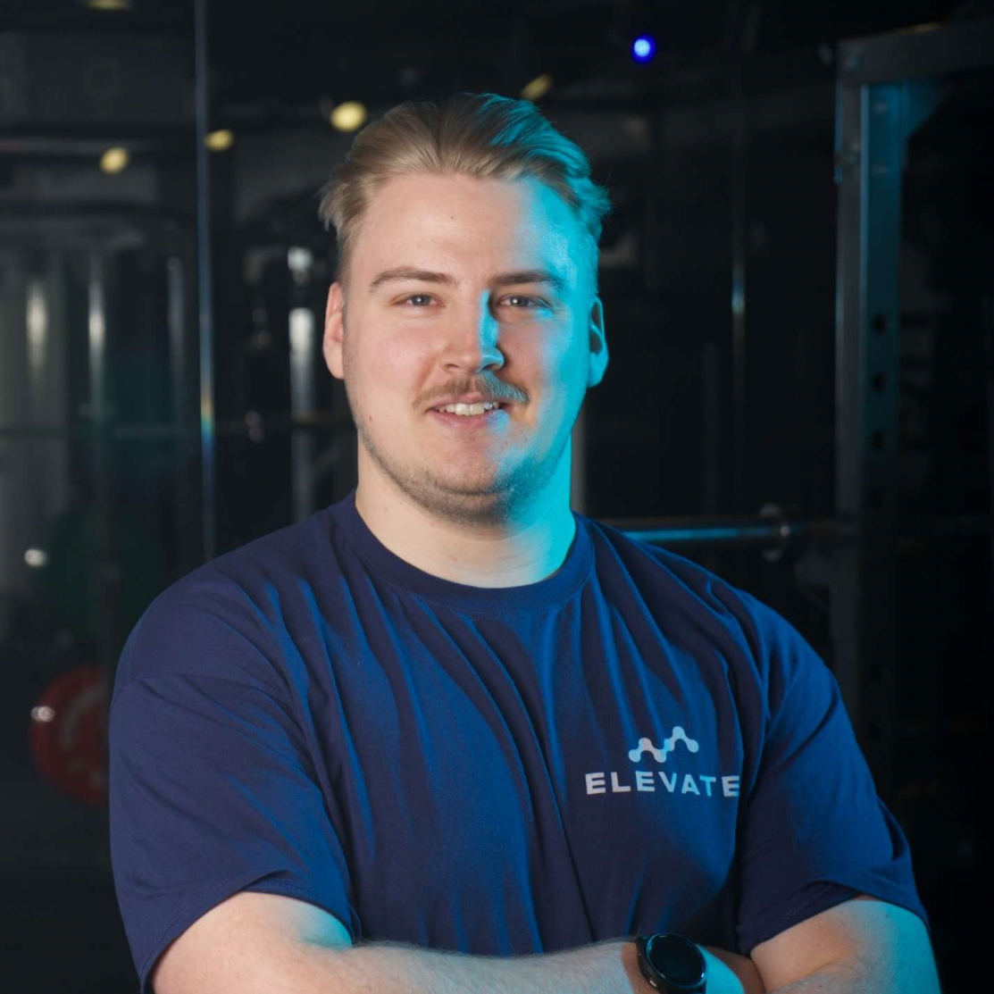 Dirk Dugdale- Duwell, wearing a blue 'Elevate' t-shirt in a dark room with gym equipment.